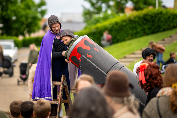 Et barn stikker hovedet op en falsk kanon, mens en voksen med kappe hjælper til.